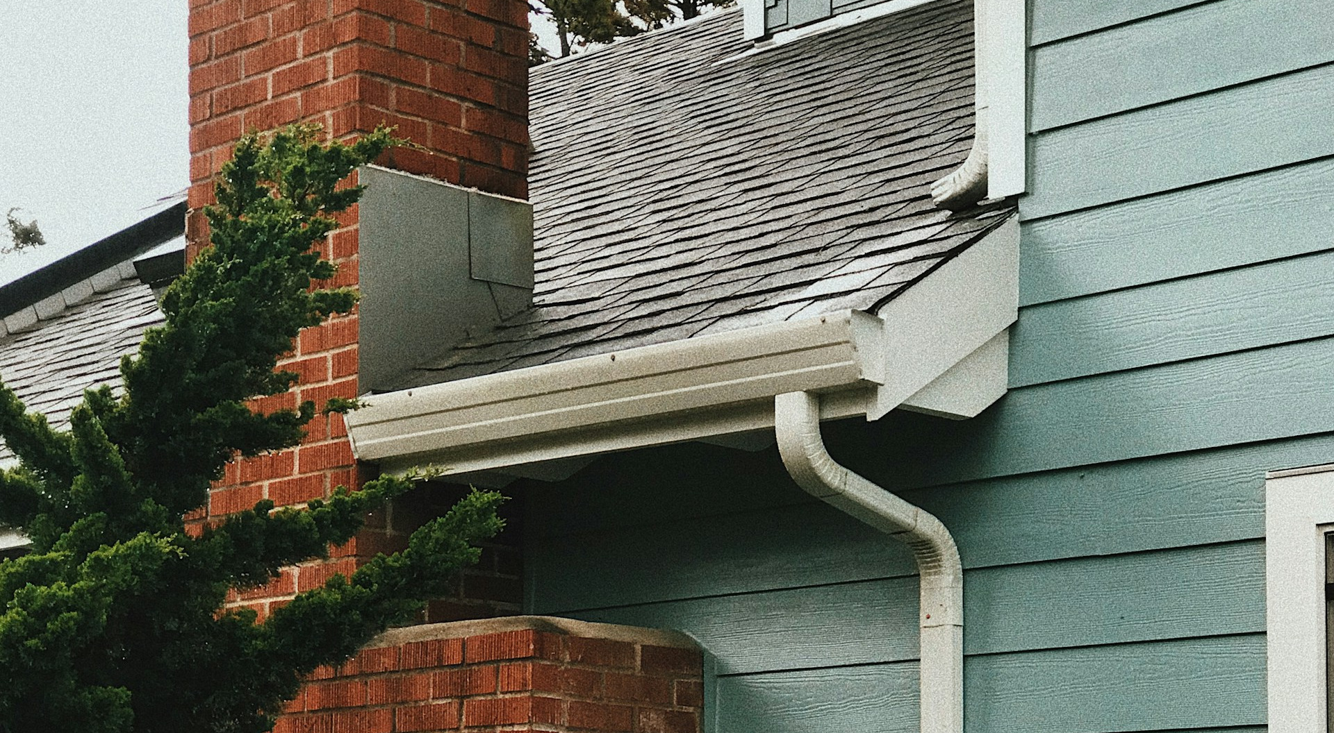 Close-up view of residential roofing materials including brick chimney, asphalt shingles, white gutters, and teal siding installation.